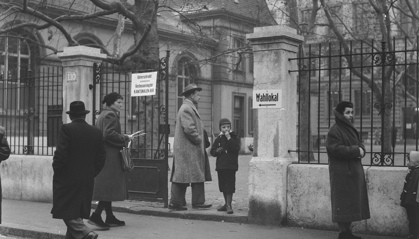 Mehrere Menschen befinden sich vor dem Hof eines Schulhauses und Wahllokals.